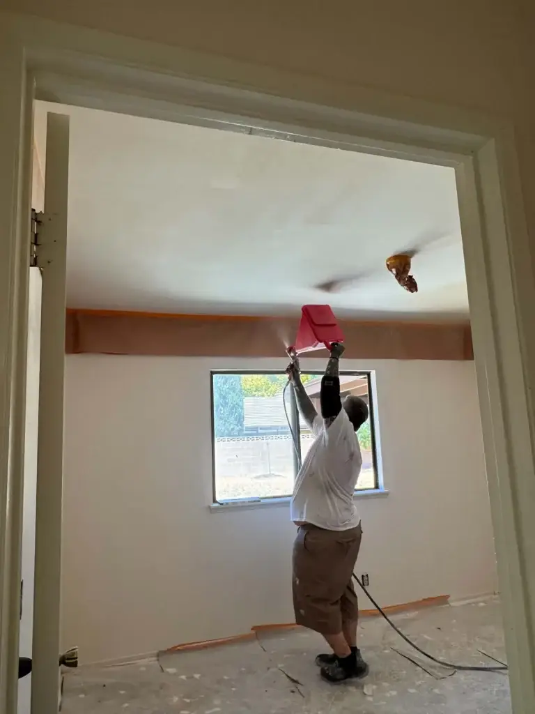 Drywall worker spraying new ceiling texture with a hopper gun in a masked-off room