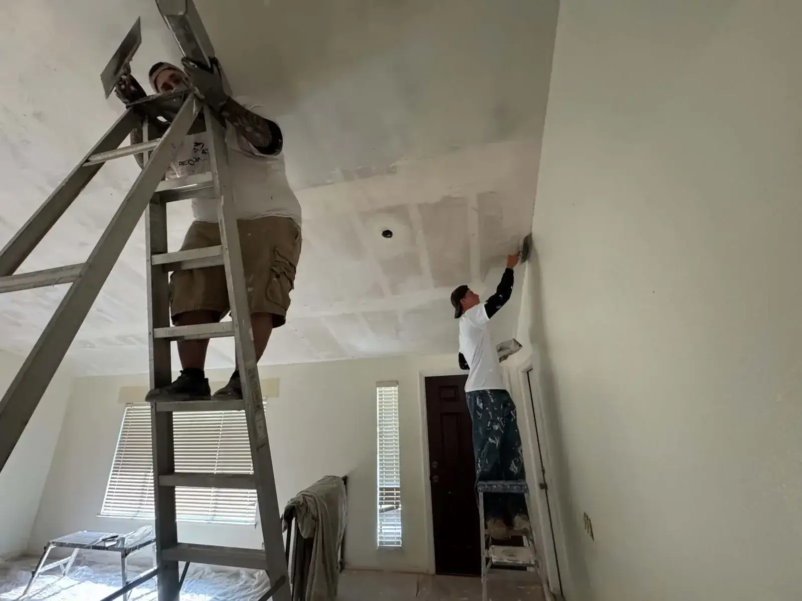 Two drywall finishers on ladders coating a ceiling with joint compound