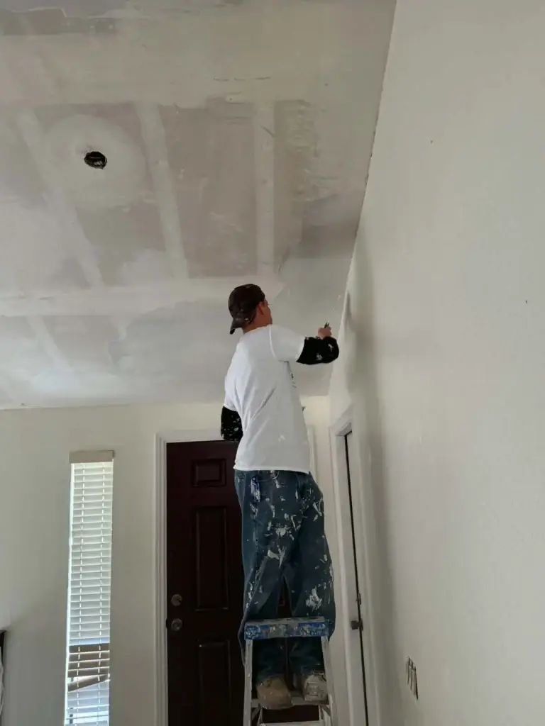 Worker finishing taped drywall seams on a ceiling near a front door